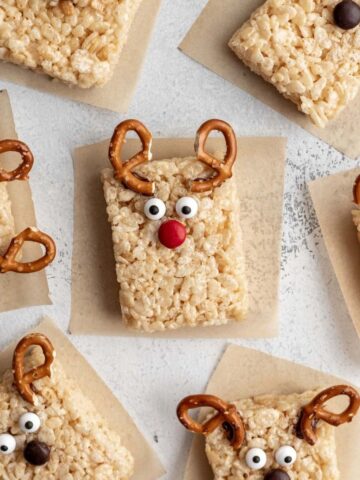 Reindeer rice krispie treats arranged on parchment paper with pretzel antlers, candy eyes, and a red candy nose.