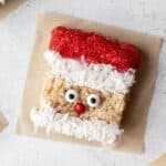 Santa rice krispie treat on parchment paper with candy eyes, red sugar hat, and a coconut beard.