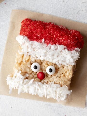 Santa rice krispie treat on parchment paper with candy eyes, red sugar hat, and a coconut beard.