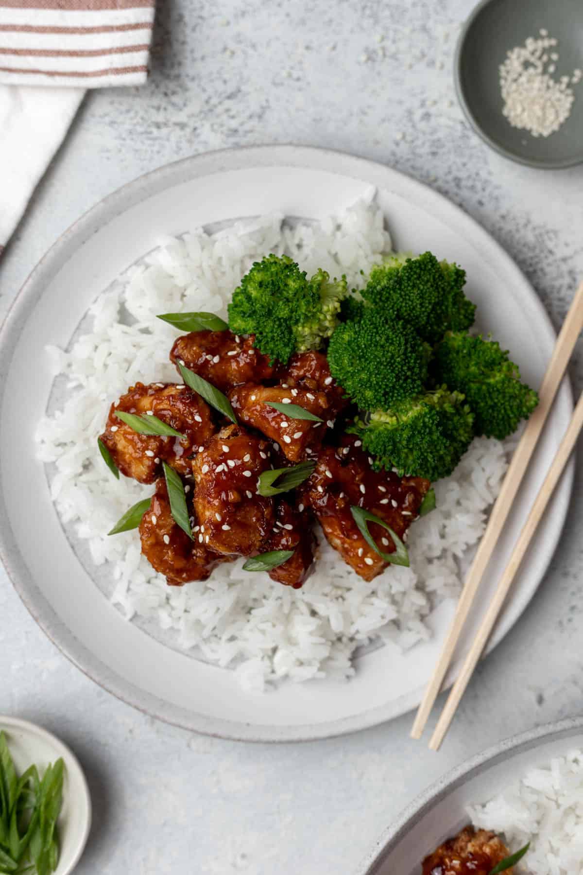 Air fryer general tso's chicken on a bed of rice with a side of steamed broccoli and chopsticks.