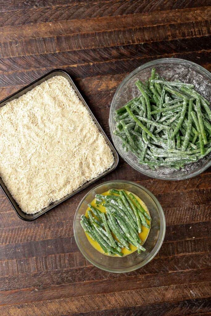 Green beans dipped in egg with a tray of breadcrumbs nearby.