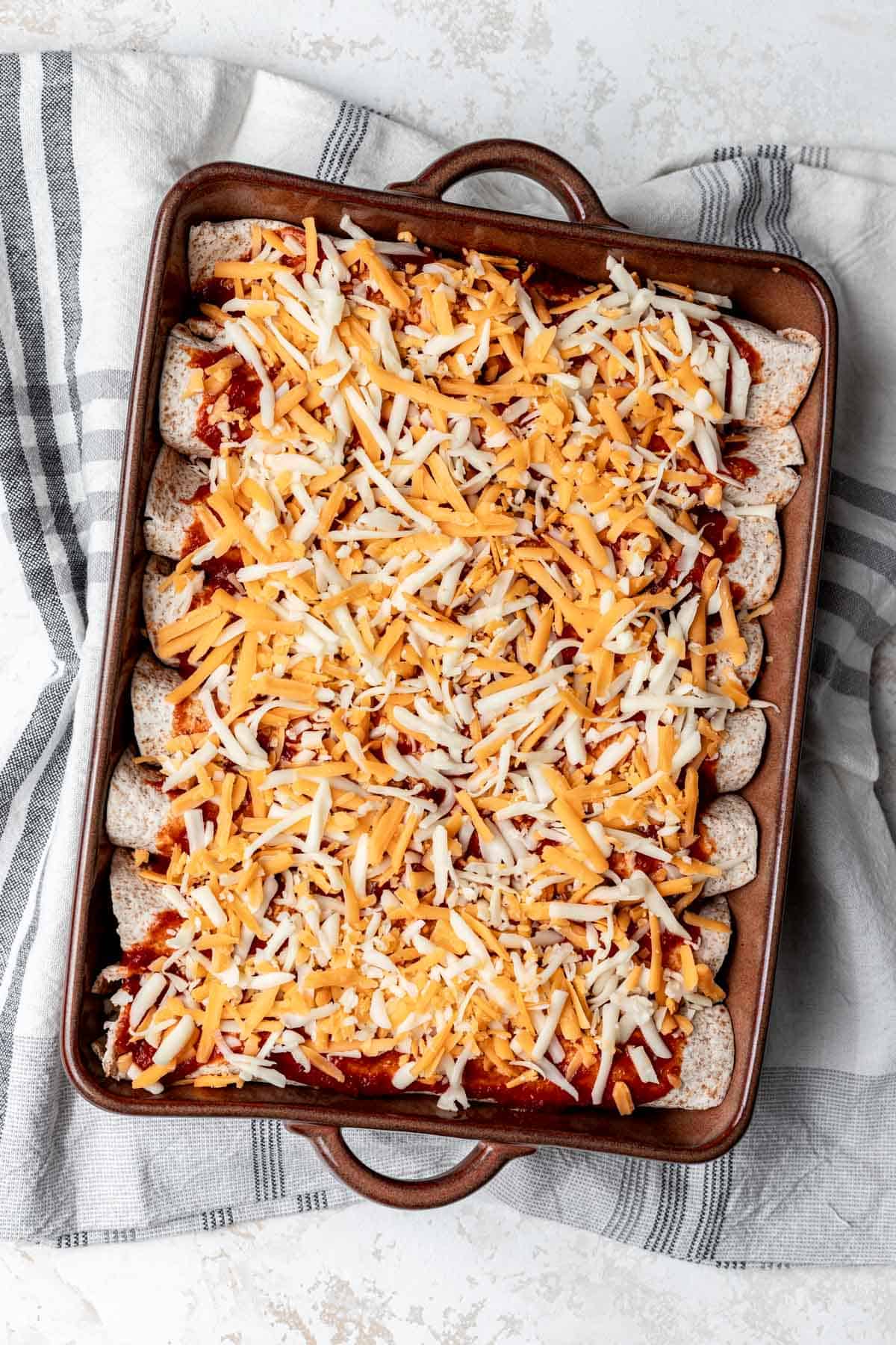 Eight rolled enchiladas topped with enchilada sauce and cheese in a baking dish.