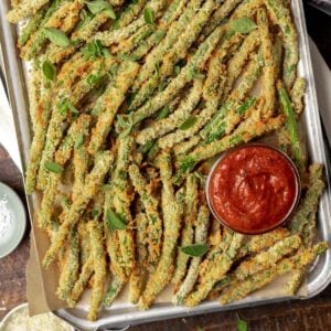 Air fryer green bean fries on a sheet tray with a side of marinara dipping sauce.