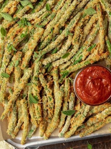 Air fryer green bean fries on a sheet tray with a side of marinara dipping sauce.