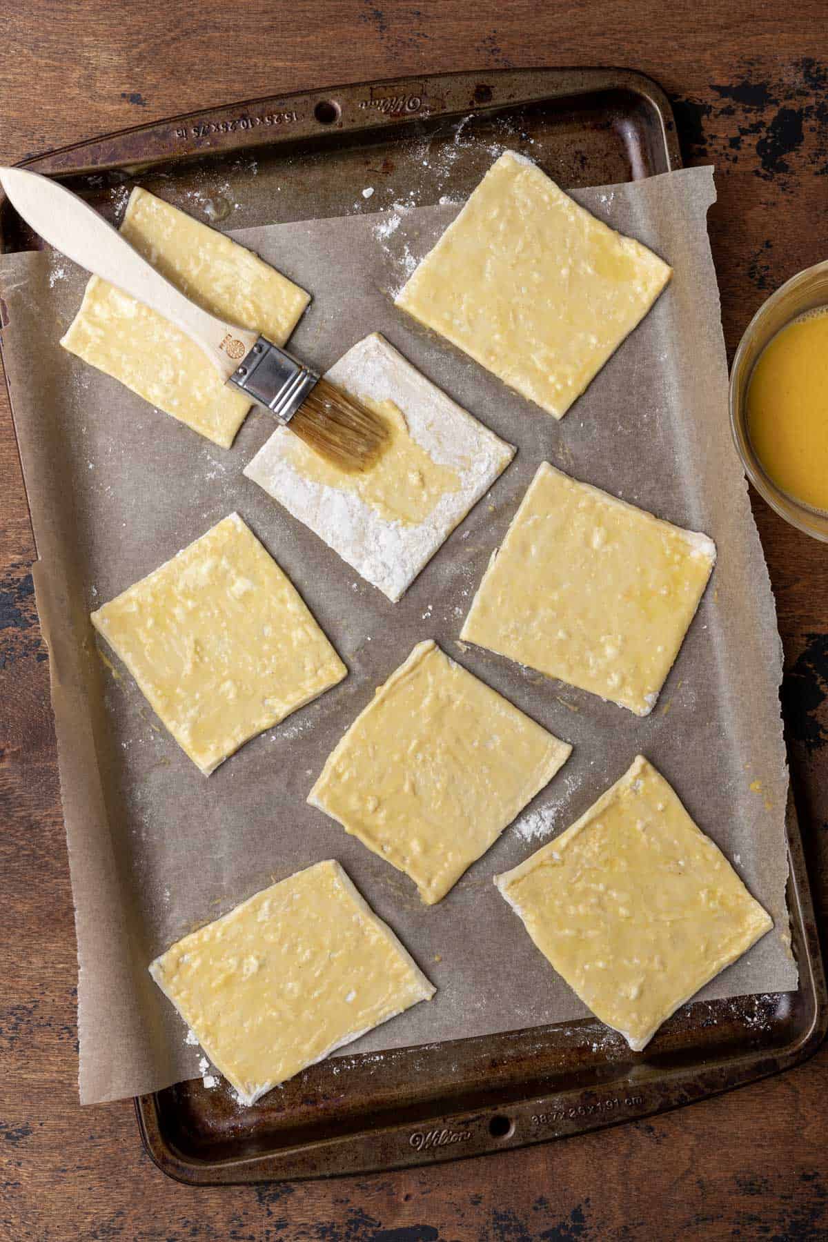 Raw squares of puff pastry dough brushed with egg on a sheet pan.
