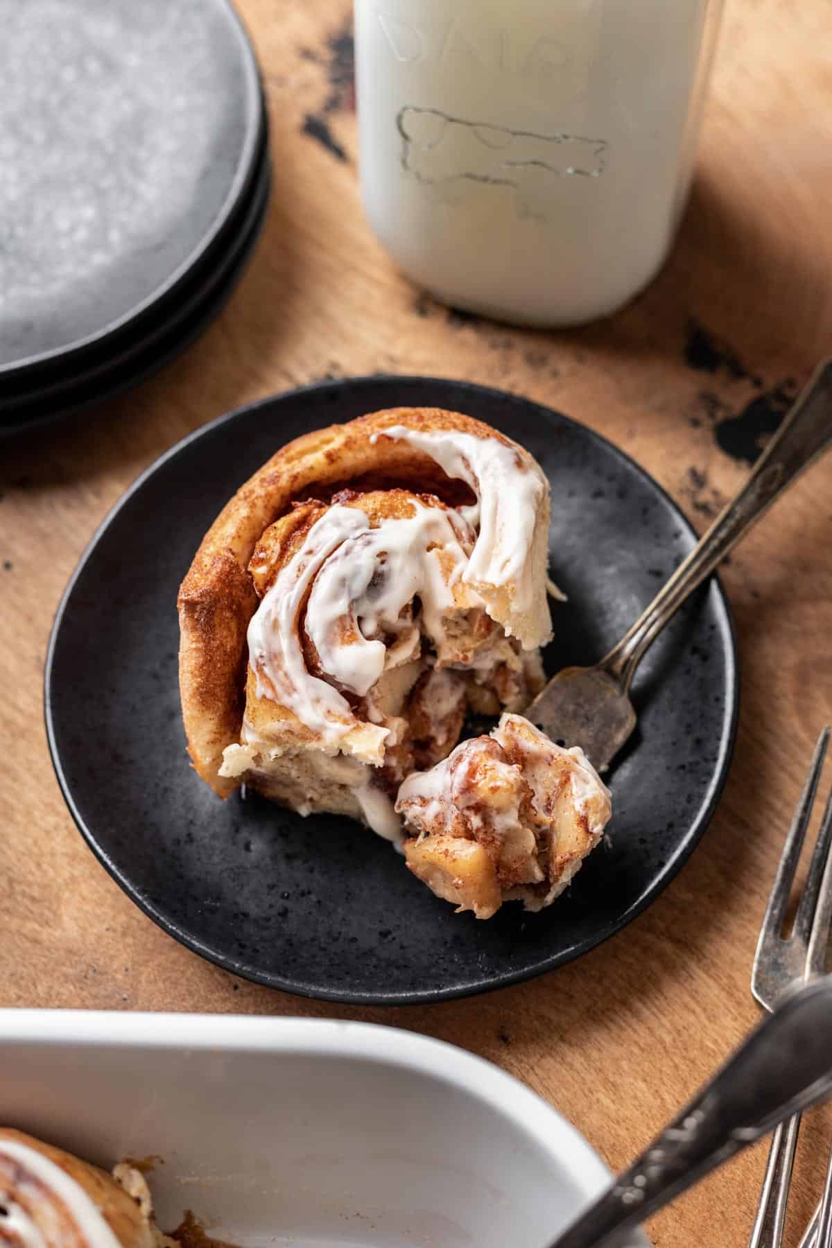 A cinnamon roll on a black plate with a fork full of apple pie filling and flakey dough.