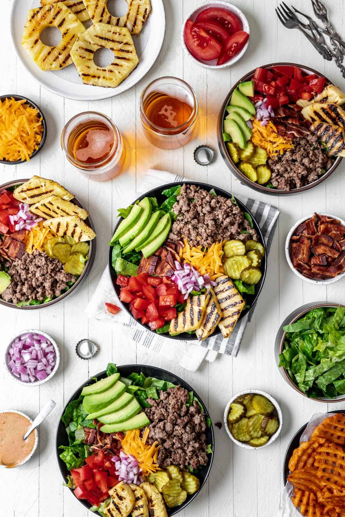 Bowls filled with lettuce, ground beef, bacon, cheese, pickles, onions, tomatoes, grilled pineapple, and avocado surrounded by the rest of the burger fixings.
