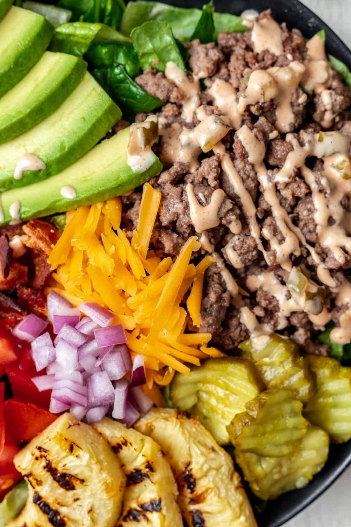 A close up view of all the burger toppings, including ground beef, sauce, pickles, cheese, grilled pineapple, and onions.