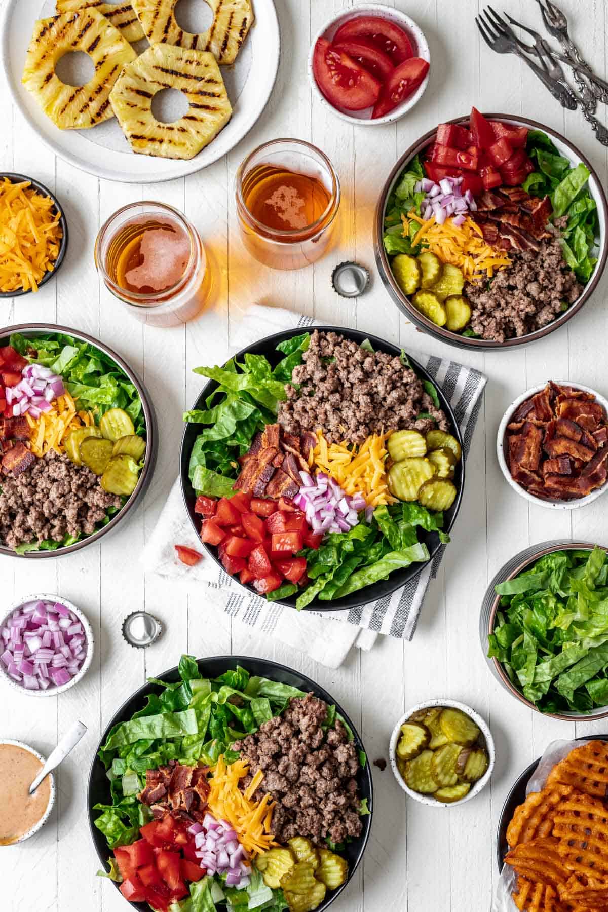Bowls filled with lettuce, ground beef, bacon, cheese, onions, pickles, and tomatoes surrounded by the rest of the burger fixings.