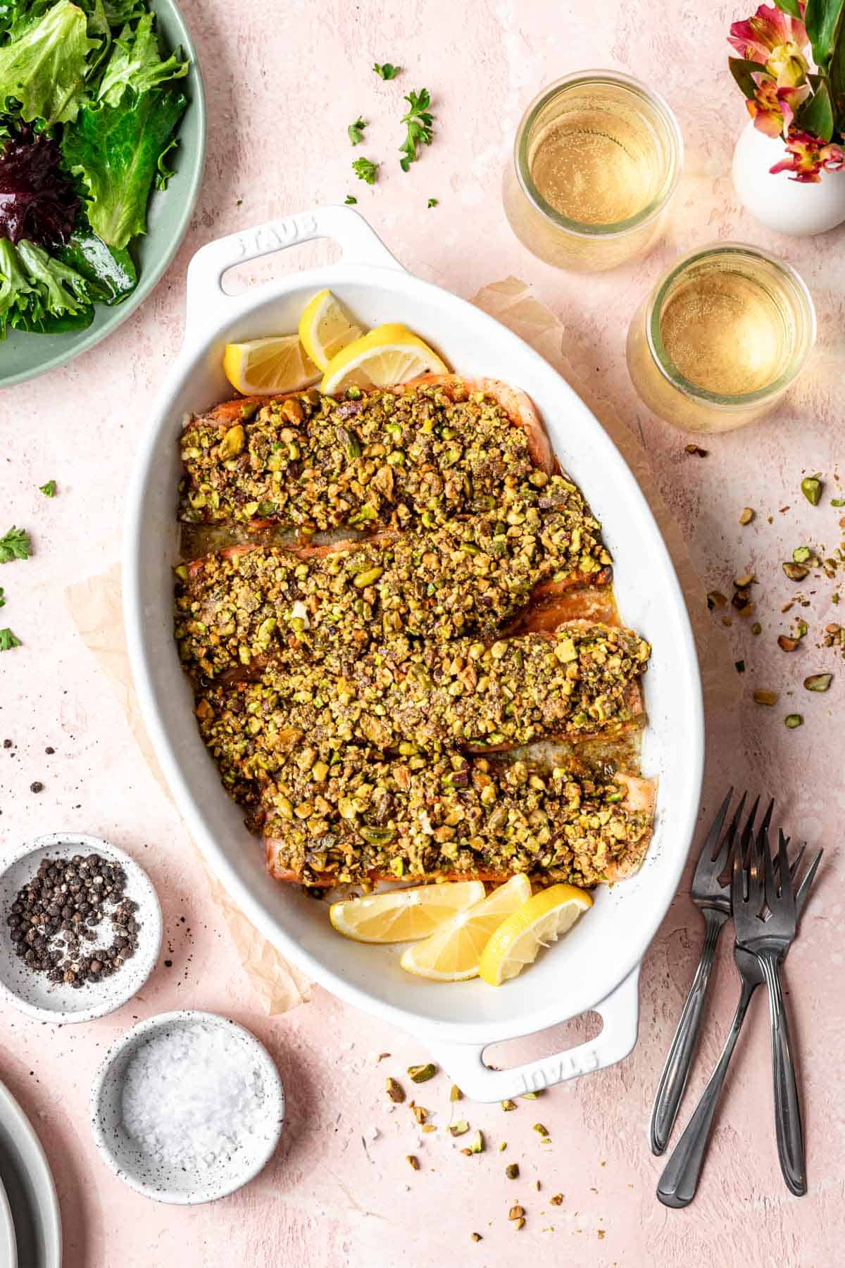4 filets of pistachio crusted salmon in a white baking dish with lemon wedges.