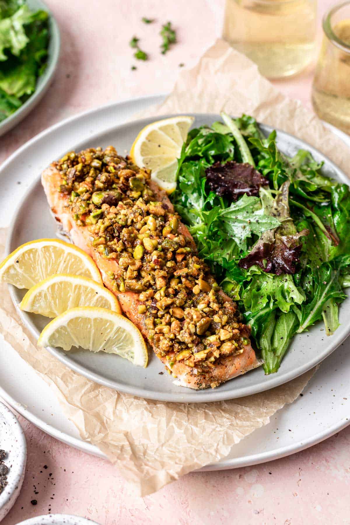 A filet of pistachio crusted salmon on a plate with a side salad and lemon wedges.