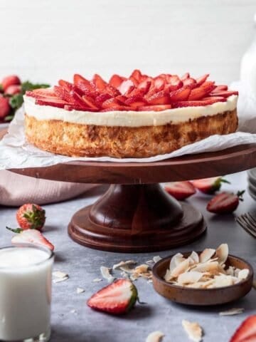 Kosher for passover cheesecake on a cake stand with toasted coconut and strawberries around it.
