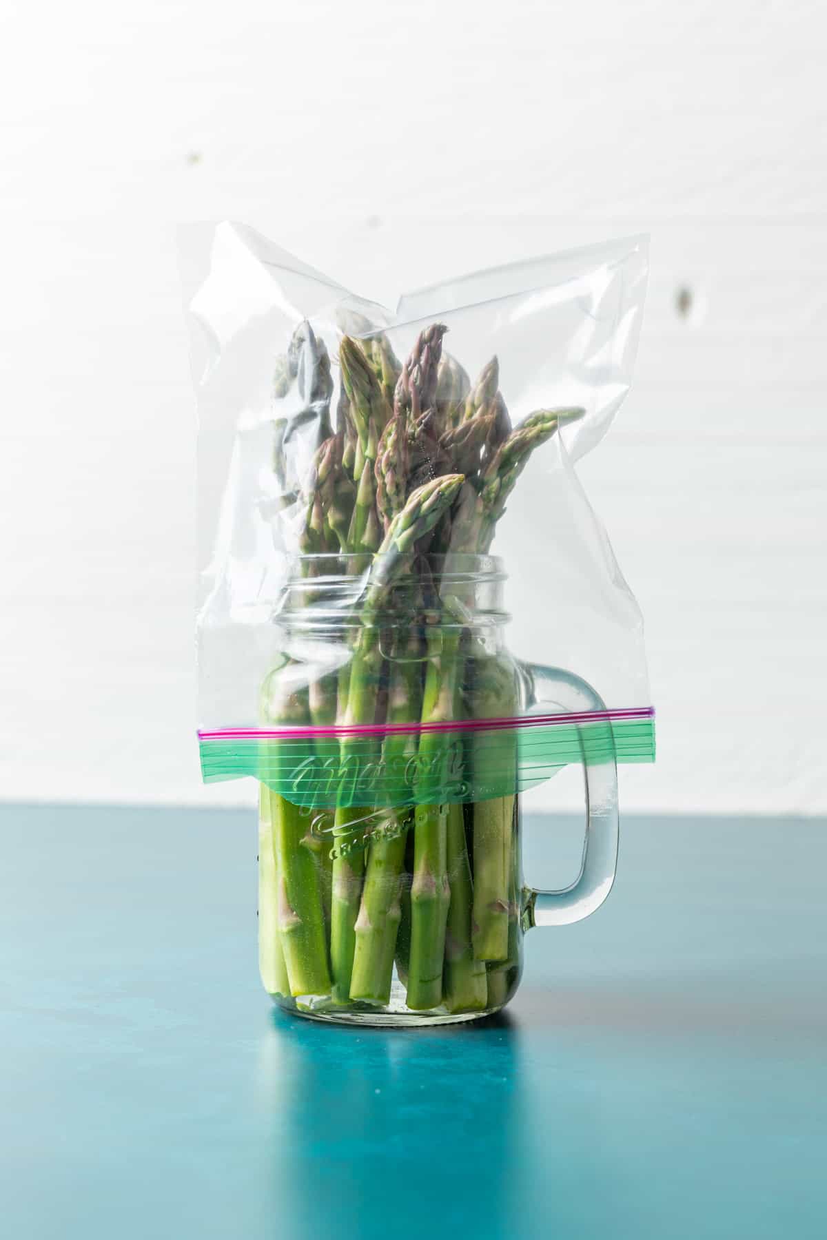 Asparagus stored in a glass jar with water and a plastic bag on top.
