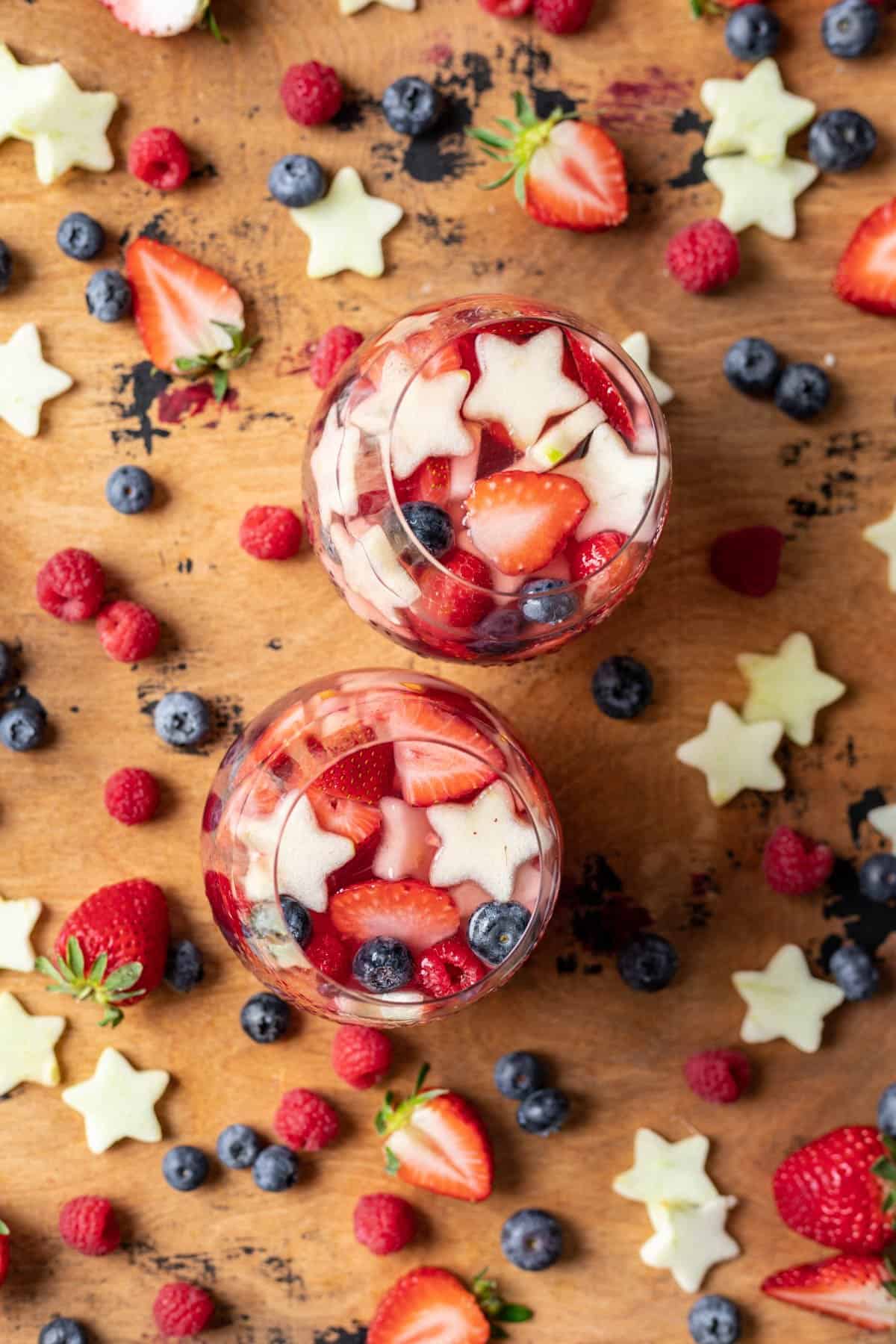 Red, white, and blue sangria in two wine glasses with fruit scattered around them.