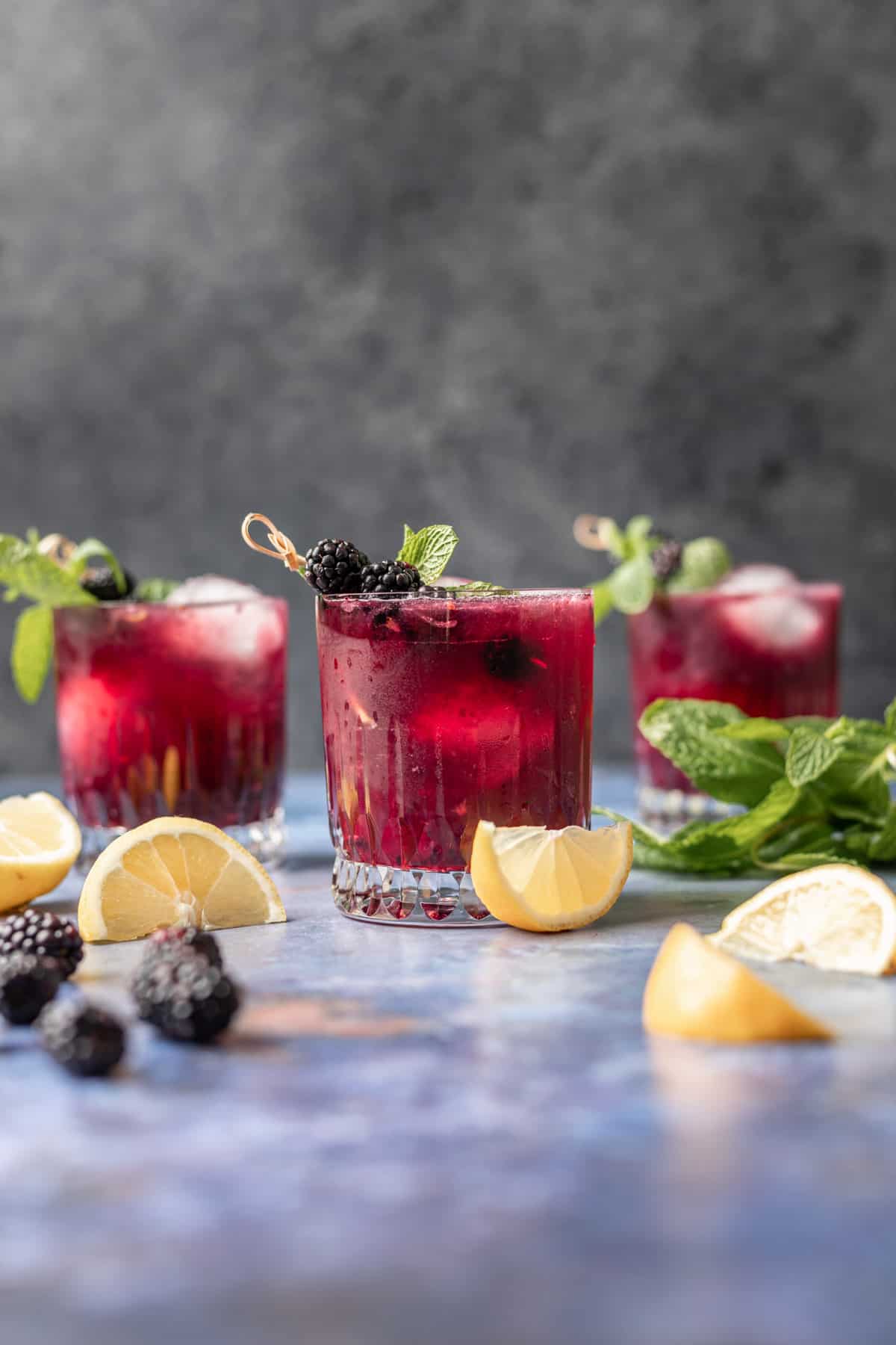 3 blackberry bourbon smash drinks surrounded by lemon wedges, fresh mint, and blackberries.