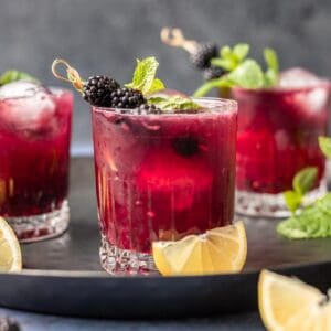3 blackberry bourbon smash drinks on a serving tray with lemon wedges.