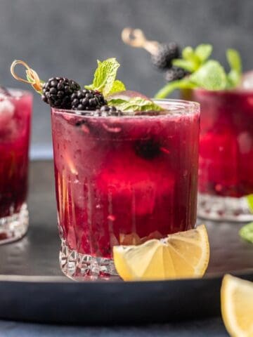 3 blackberry bourbon smash drinks on a serving tray with lemon wedges.