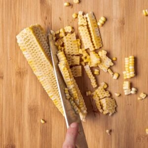 A knife cutting corn off of a cob onto a cutting board.