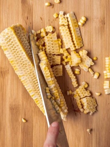 A knife cutting corn off of a cob onto a cutting board.