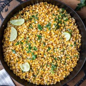 Skillet roasted corn kernels in a cast iron skillet with parsley and lime wedges.