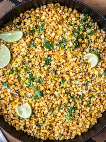 Skillet roasted corn kernels in a cast iron skillet with parsley and lime wedges.