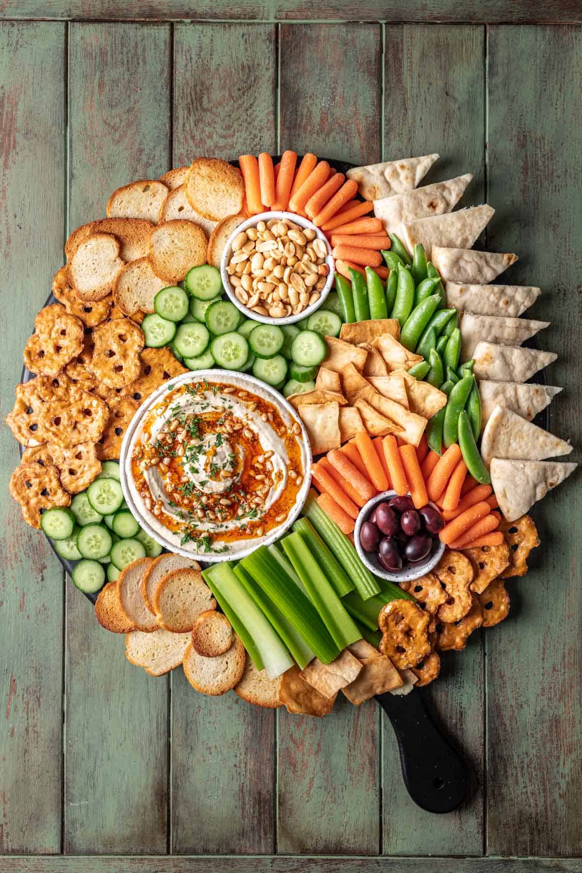 A large snack tray with hummus, vegetables, nuts, olives, and chips.