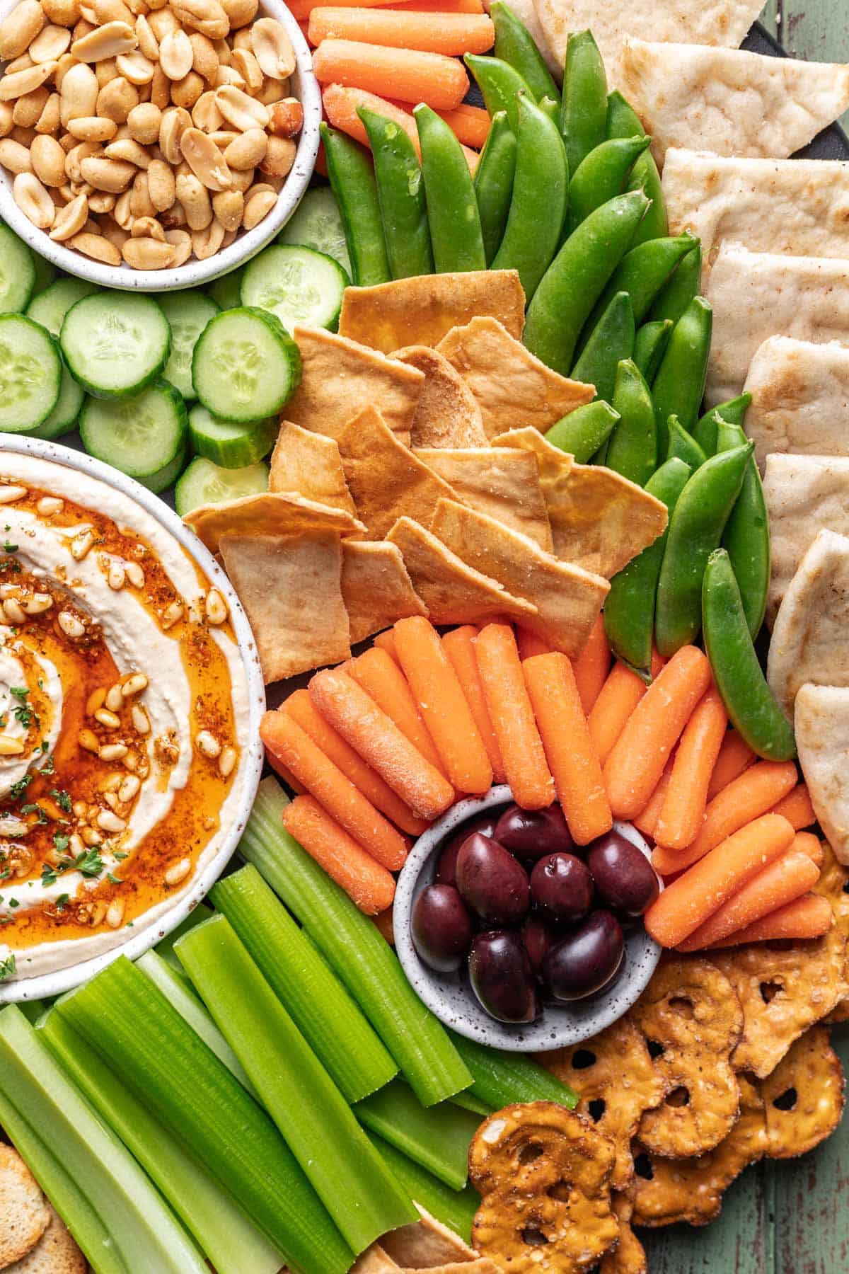 Snacks arranged on a board, like cucumbers, olives, pita chips, snap peas, and celery.