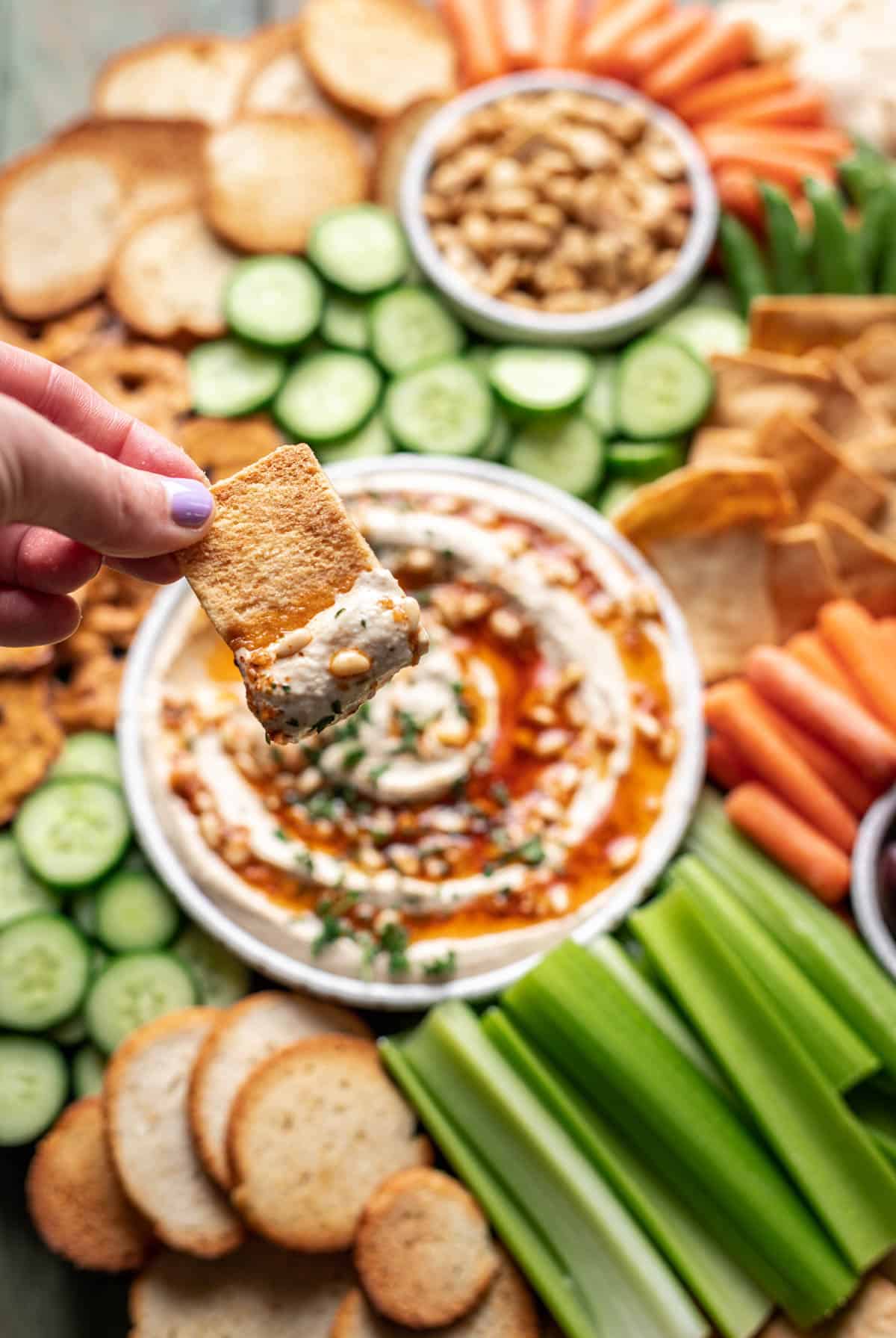 A hand dipping a pita chip into a plate of pine nut hummus.