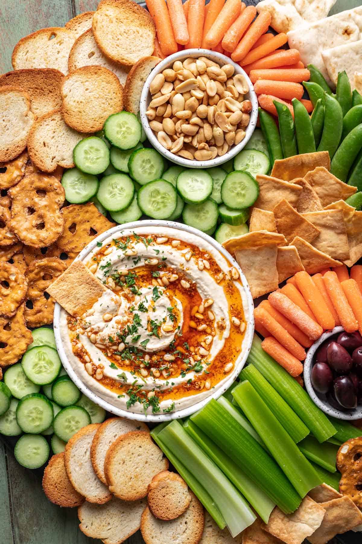 A snack board with pine nut hummus, chips, pretzels, nuts, olives, and fresh vegetables.