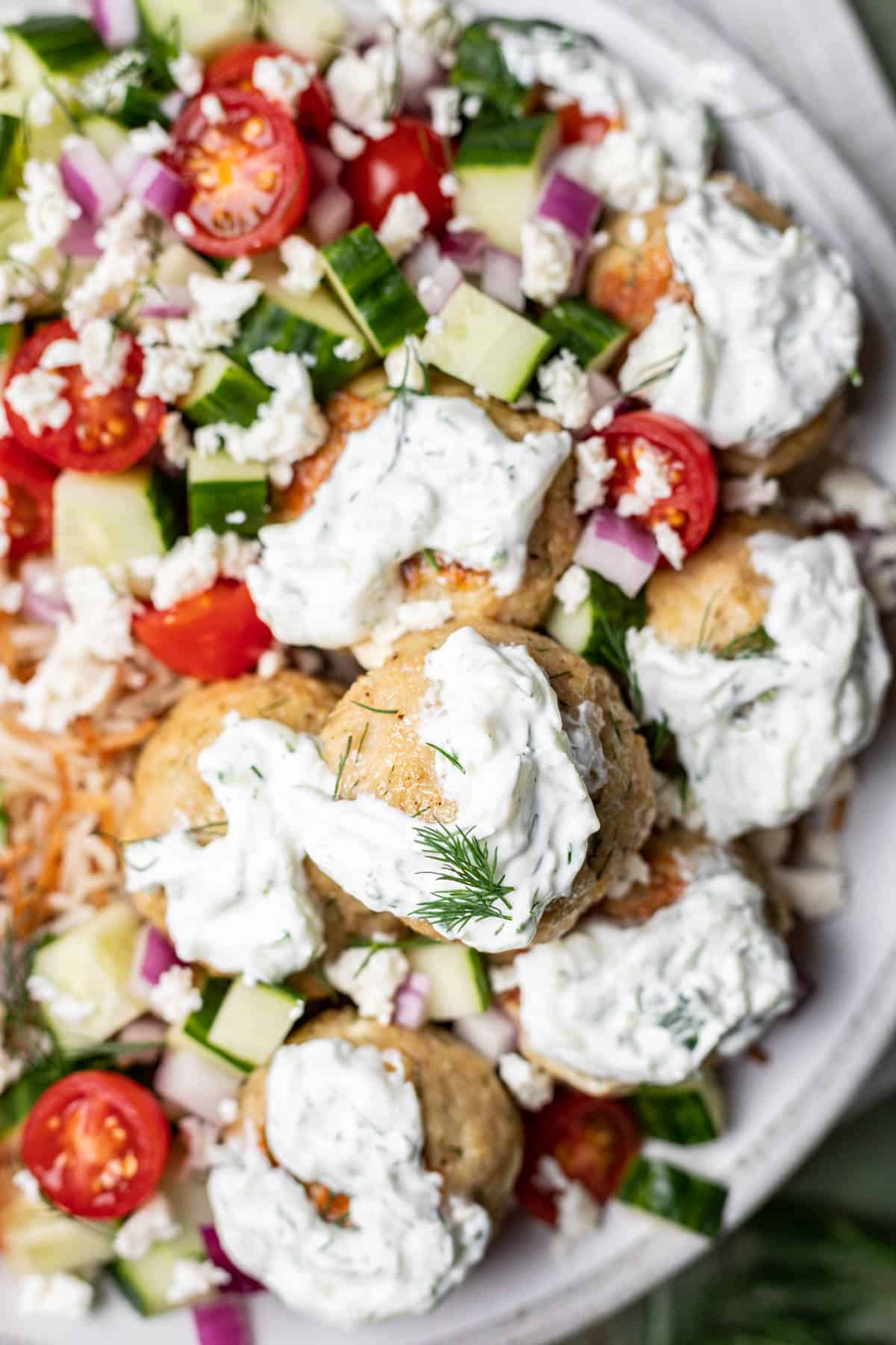 Greek chicken meatballs topped with tzatziki sauce in a bowl with cucumber, tomato, and feta.