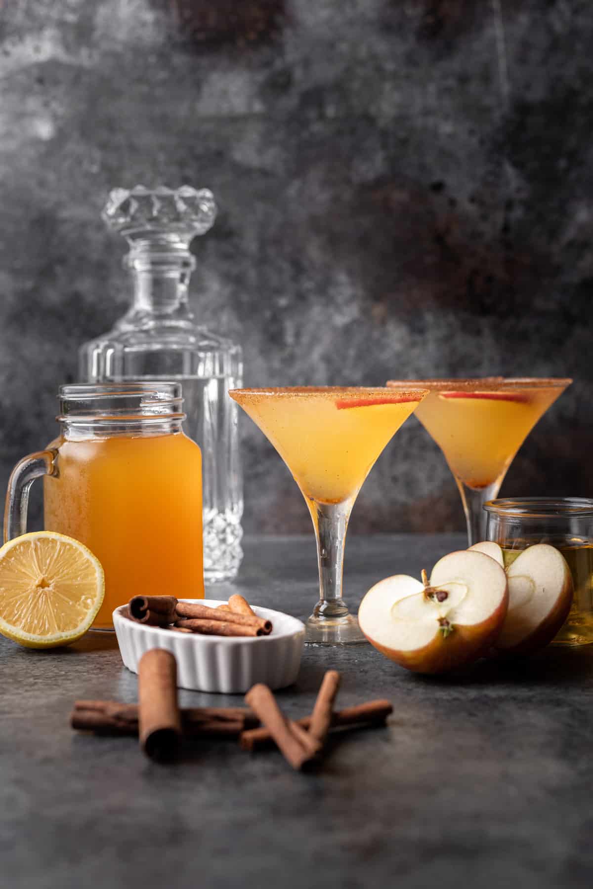 Apple cider martinis with cinnamon sticks and sliced apples next to them.