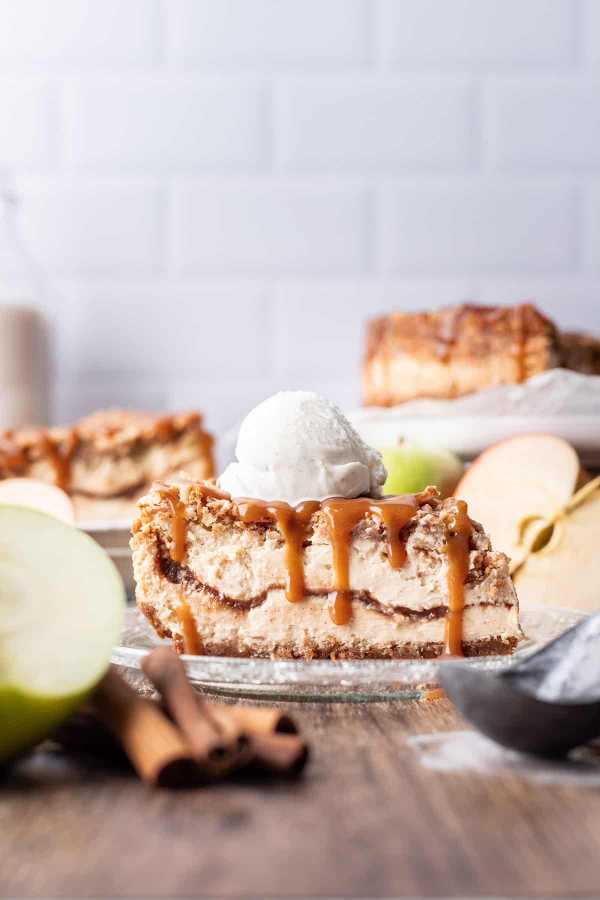 A slice of apple crumble cheesecake on a glass plate with caramel dripping down the side.