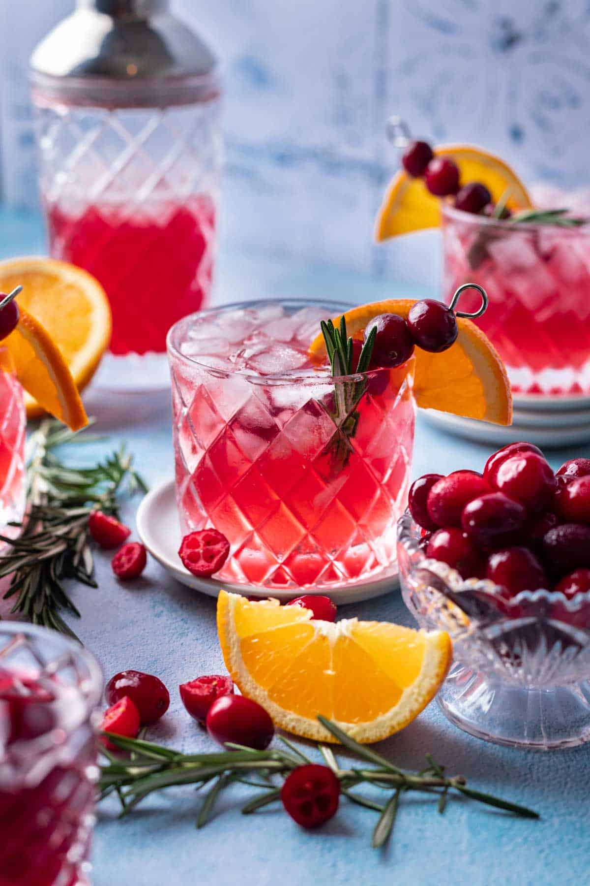 Cranberry gin cocktail on a blue backdrop with fresh cranberries and orange slices around it.