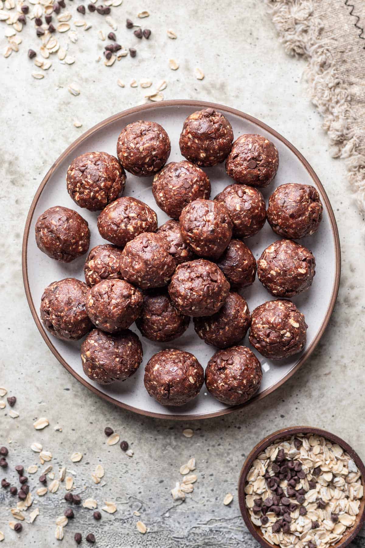 Chocolate date balls on a plate with oats and chocolate chips scattered around it.