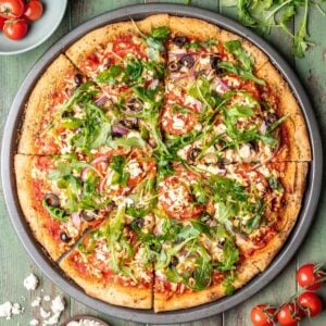 Mediterranean pizza on a pizza pan surrounded with tomatoes, arugula, and feta.