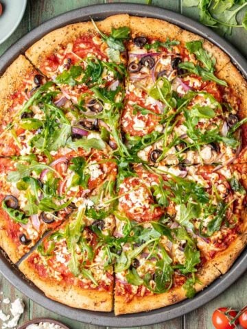 Mediterranean pizza on a pizza pan surrounded with tomatoes, arugula, and feta.