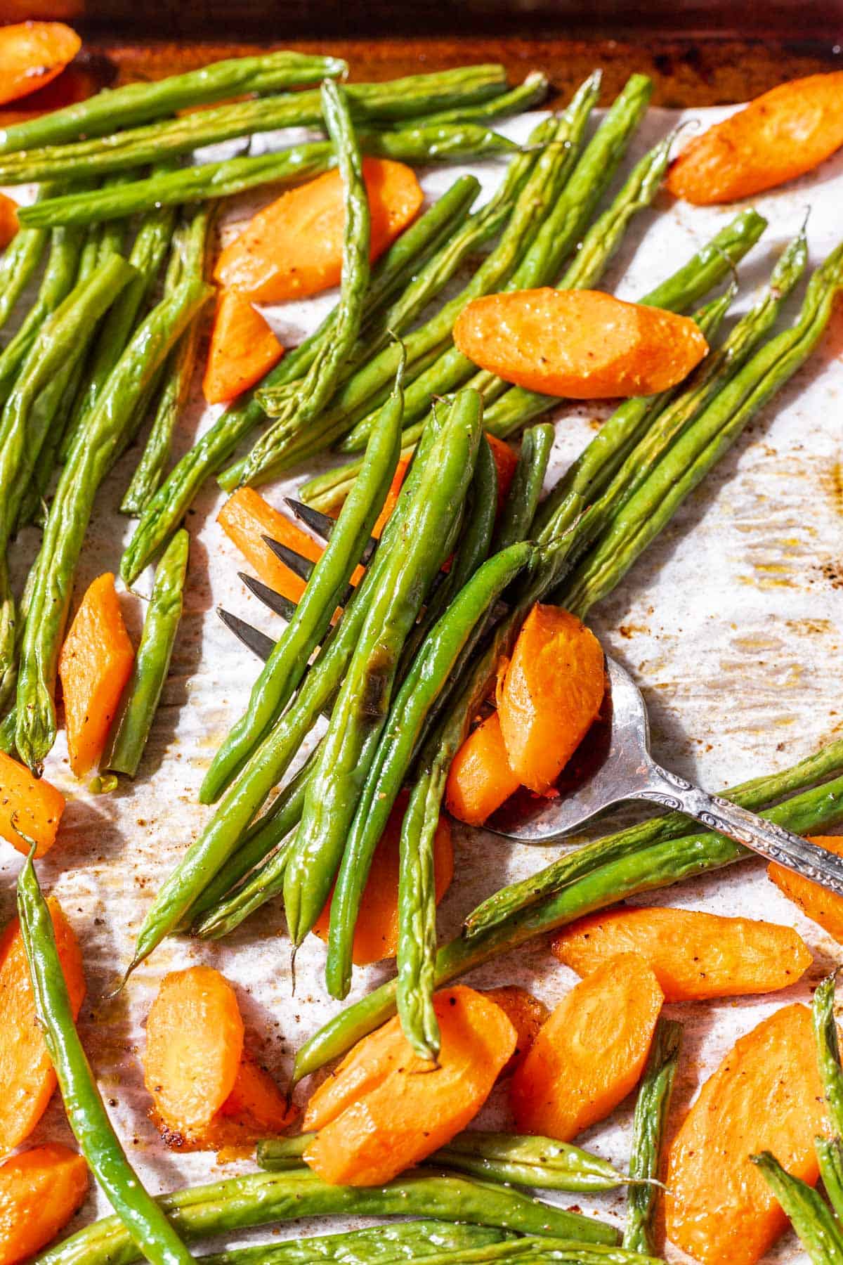 A serving fork scooping roasted green beans and carrots off a baking sheet.