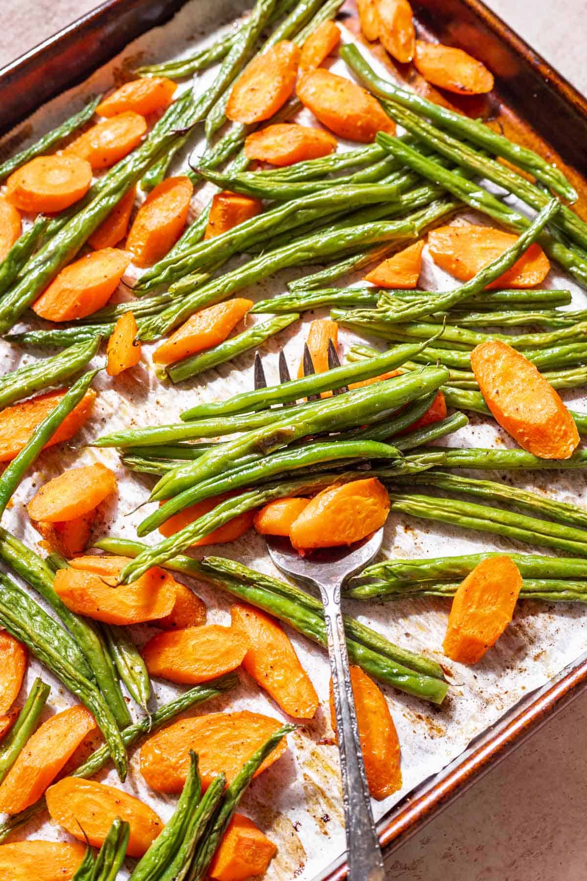 Roasted green beans and carrots on a baking sheet with a serving fork.