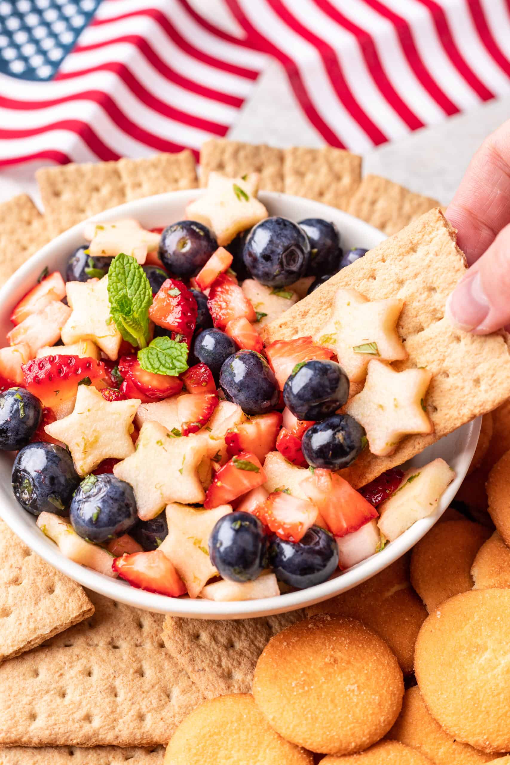 A hand scooping a graham cracker into a bowl of red, white, and blue fruit salsa.