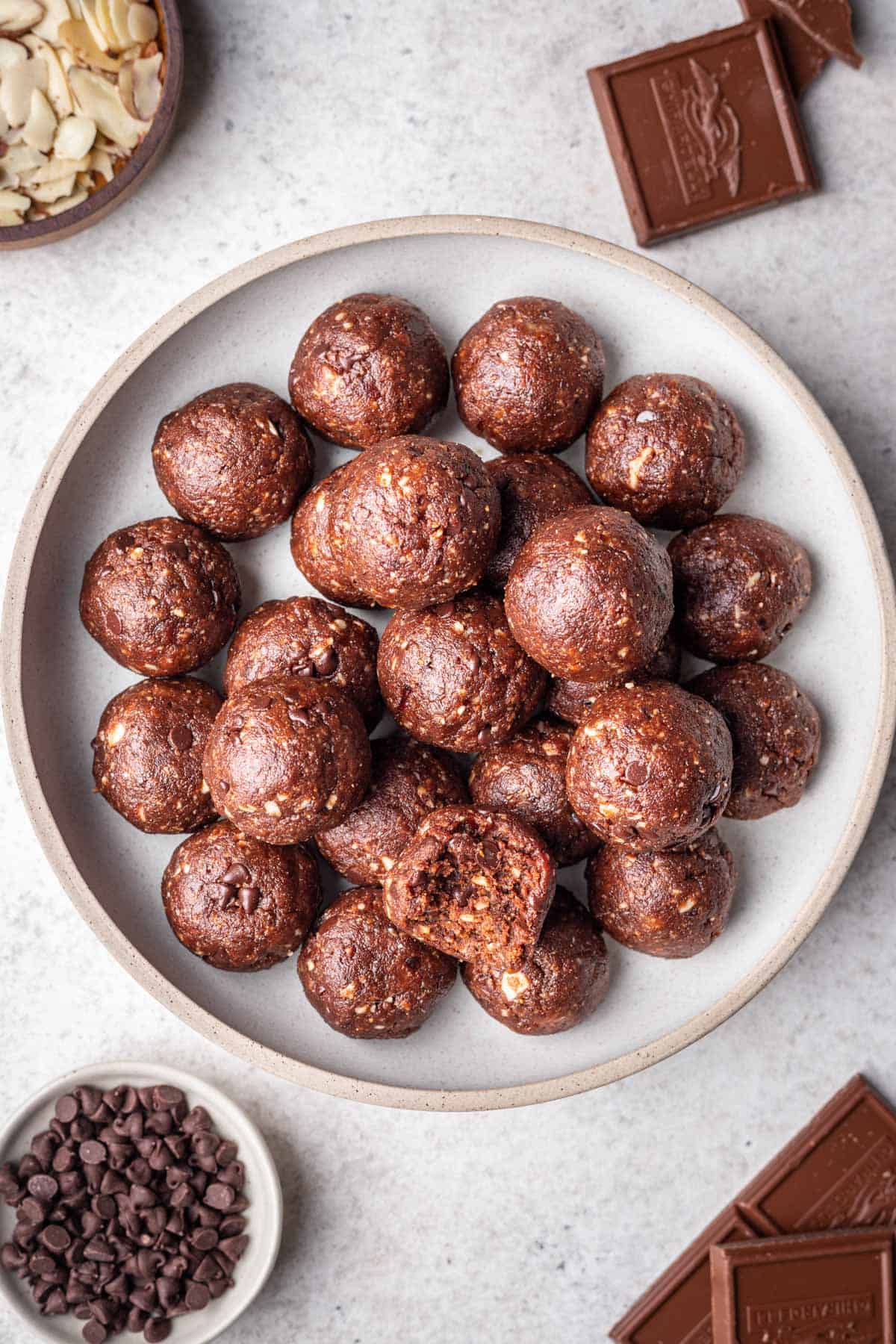 Chocolate bliss balls in a serving bowl surrounded by chocolate chips, chocolate bars, and sliced almonds.