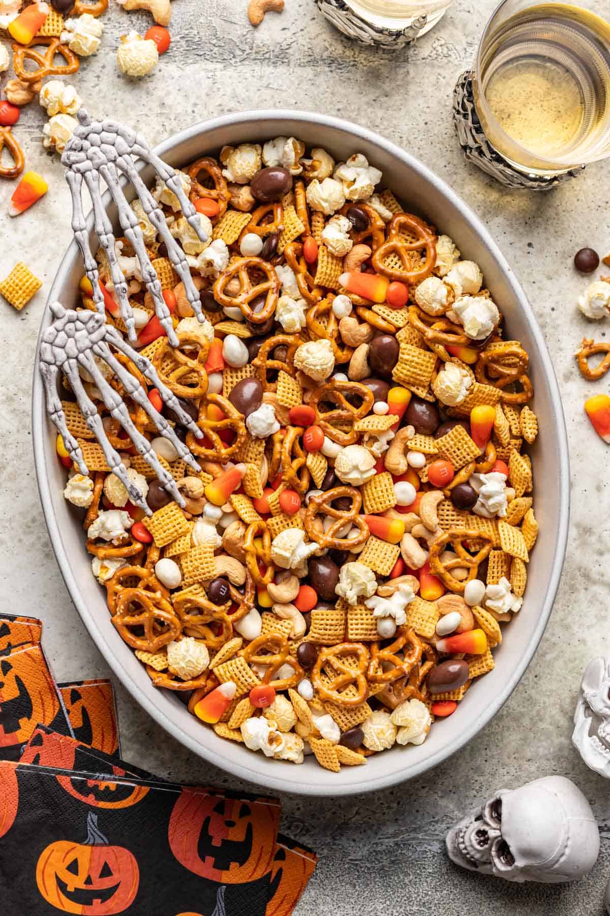 Halloween trail mix surrounded by drinks, pumpkin decorated napkins, and decorative skeleton skulls.
