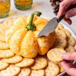 A cheese knife cutting into a pumpkin shaped cheese ball.