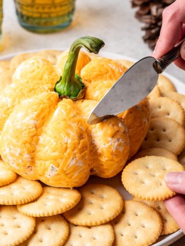 A cheese knife cutting into a pumpkin shaped cheese ball.