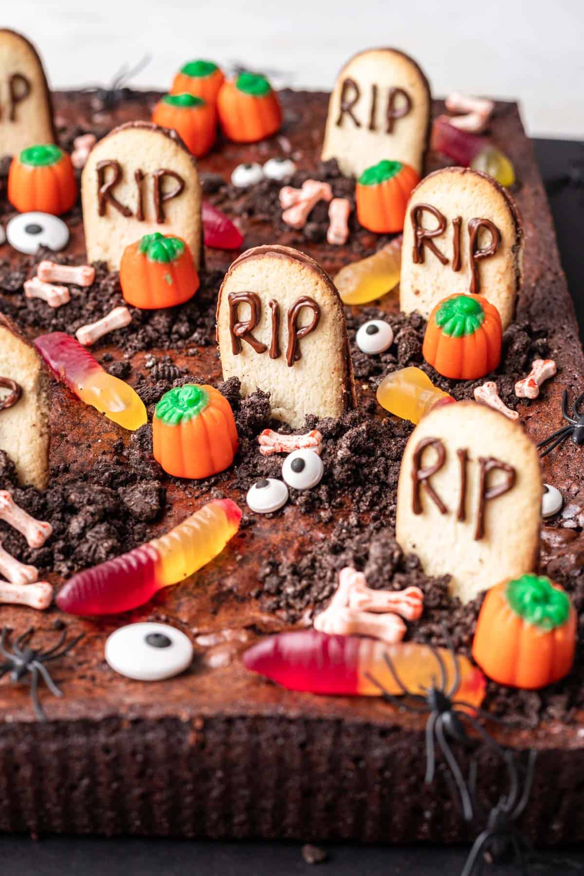 Graveyard brownies decorated with tombstone cookies, crushed oreo cookies, candy pumpkins, and gummy worms.