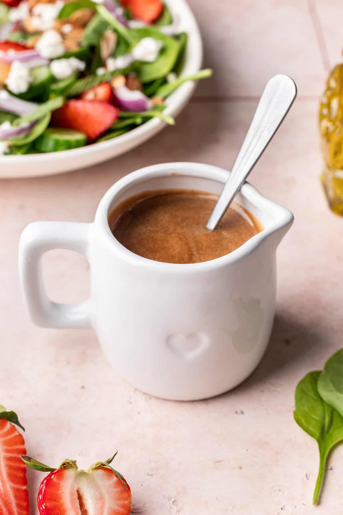 Creamy maple balsamic dressing in a glass container with a salad, oil, and leafy greens around it.