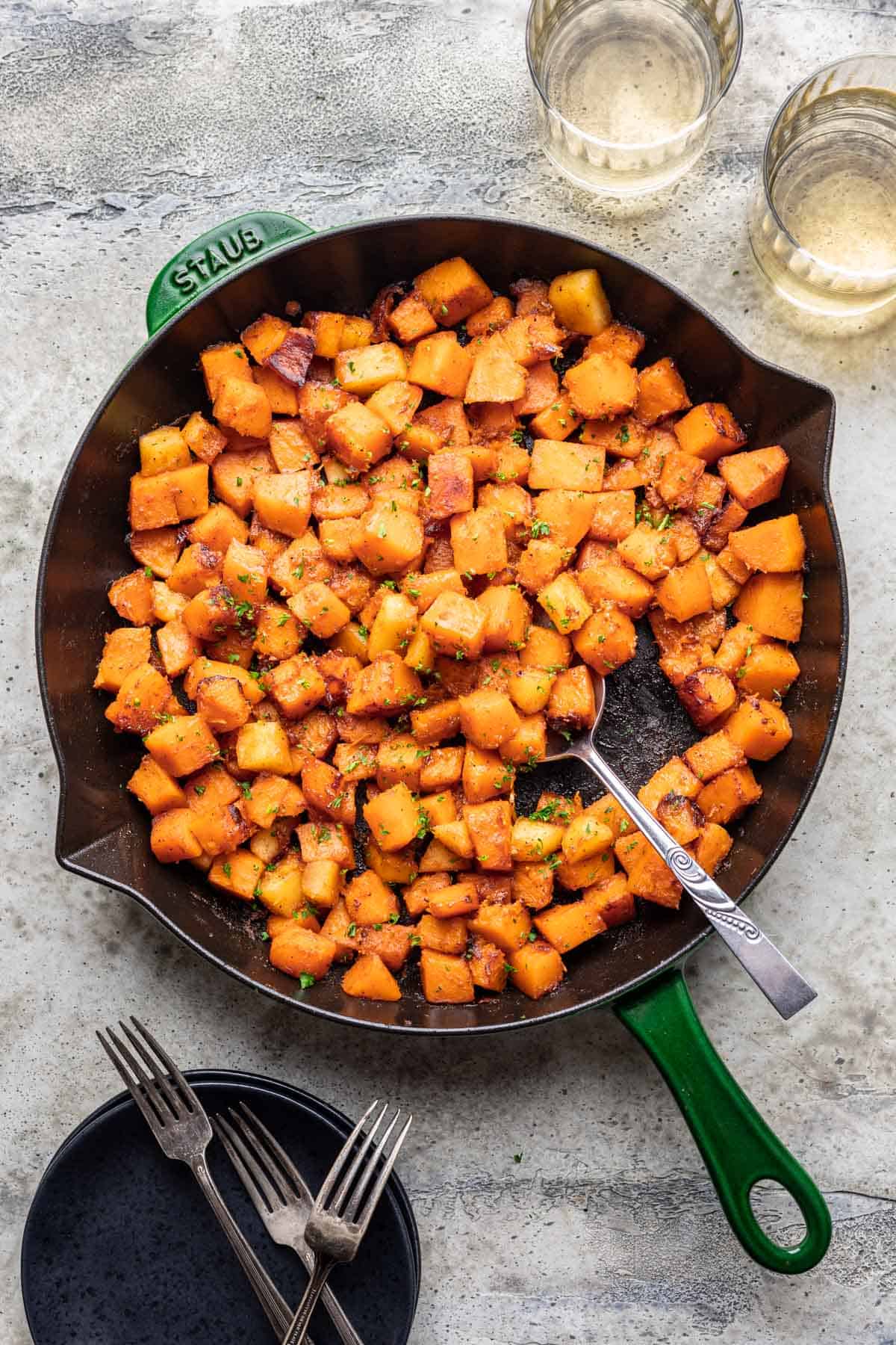 Sautéed butternut squash in a skillet with serving plates and glasses of wine next to it.