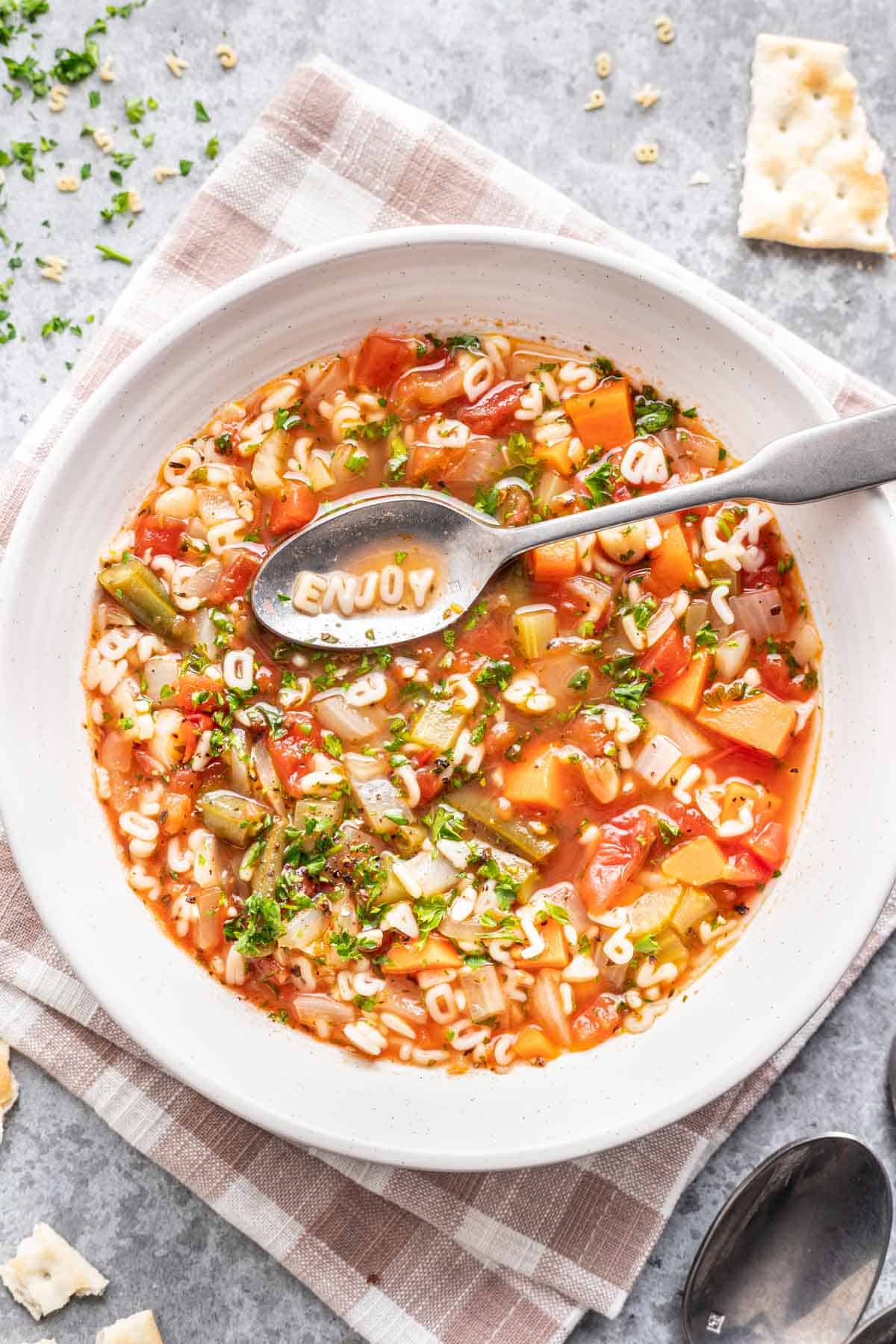 Homemade alphabet soup in a white bowl with ABC pasta spelling "ENJOY" on a spoon.