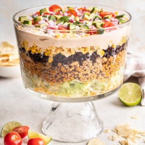 Layered taco salad in a trifle dish topped with avocado, tomatoes, and red onions.