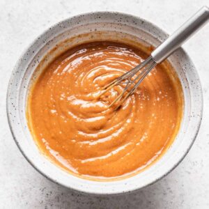 Spicy peanut sauce in a white bowl with a small whisk.