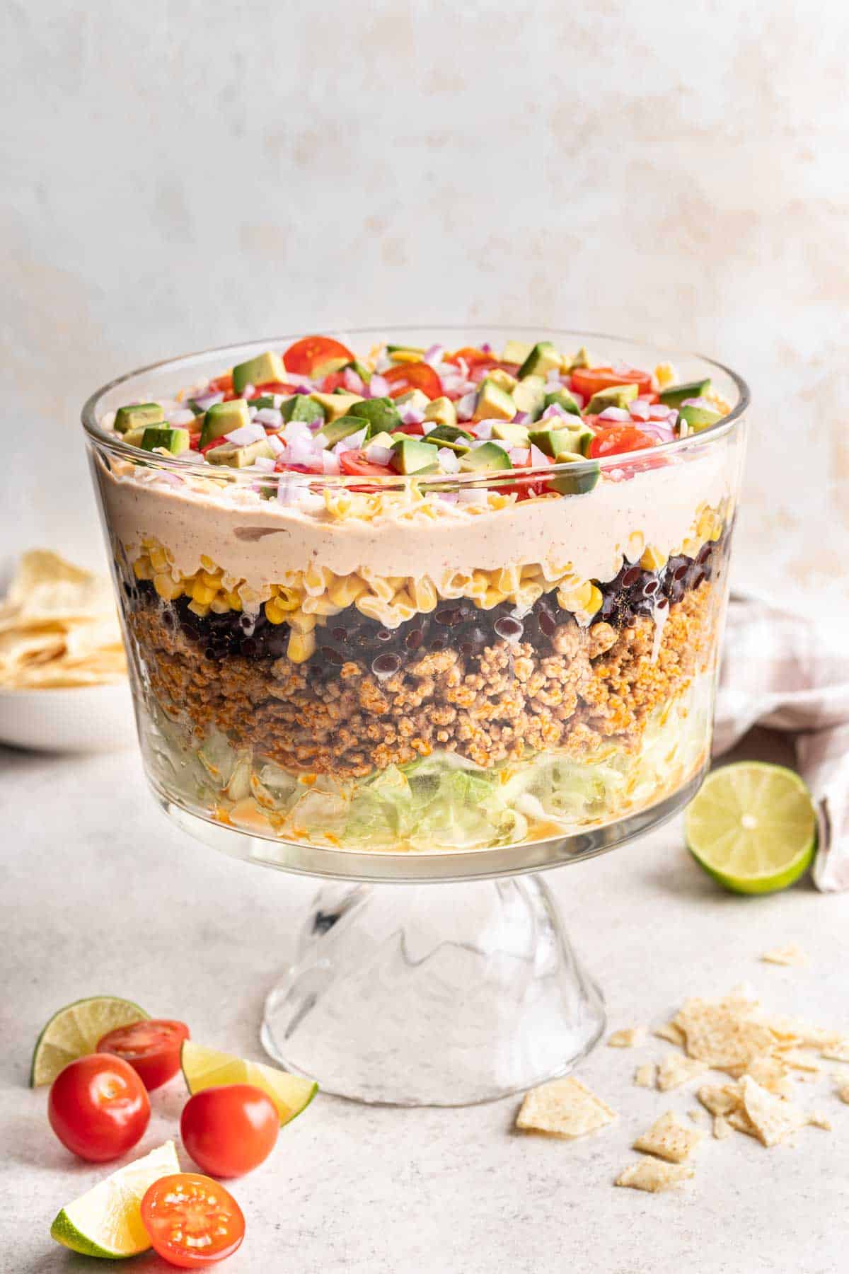 Layered taco salad in a trifle dish with tomatoes, avocado, and onions on top.